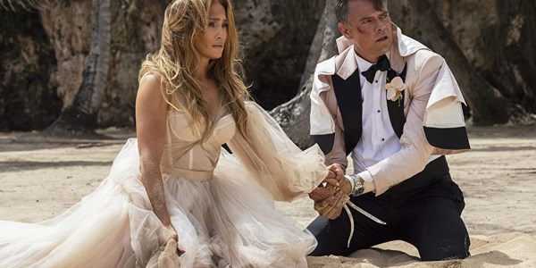 Jennifer Lopez and Josh Duhamel in Shotgun Wedding. They're kneeling on the beach, hands tied together, covered in dirt and blood, after their wedding was ambushed by armed pirates.
